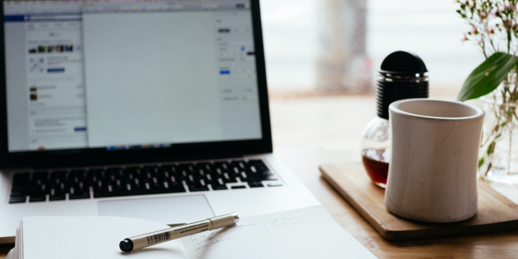 Laptop on a desk with a notebook in front and a pen on top. There is a mug and coffee to the right of the laptop.