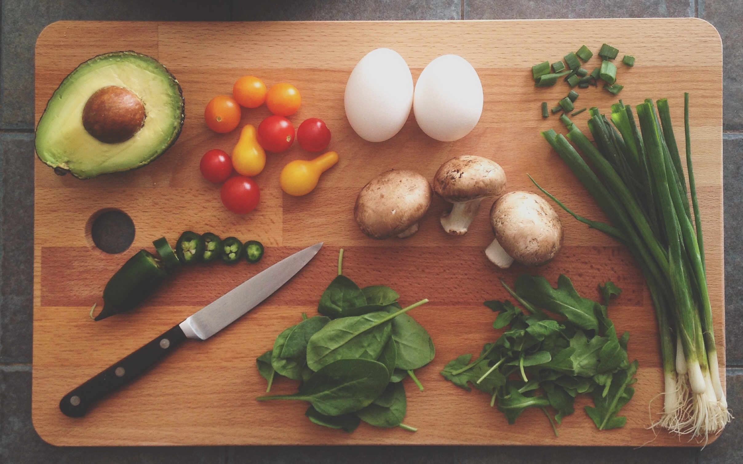 Wooden chopping board with a selection of cut up vegetables - half an avocado chilli, spinach, tomatoes, mushrooms, spring onions, salad leaves and mushrooms.